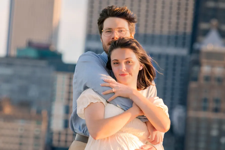He holds her from behind, both smiling, downtown towers behind them